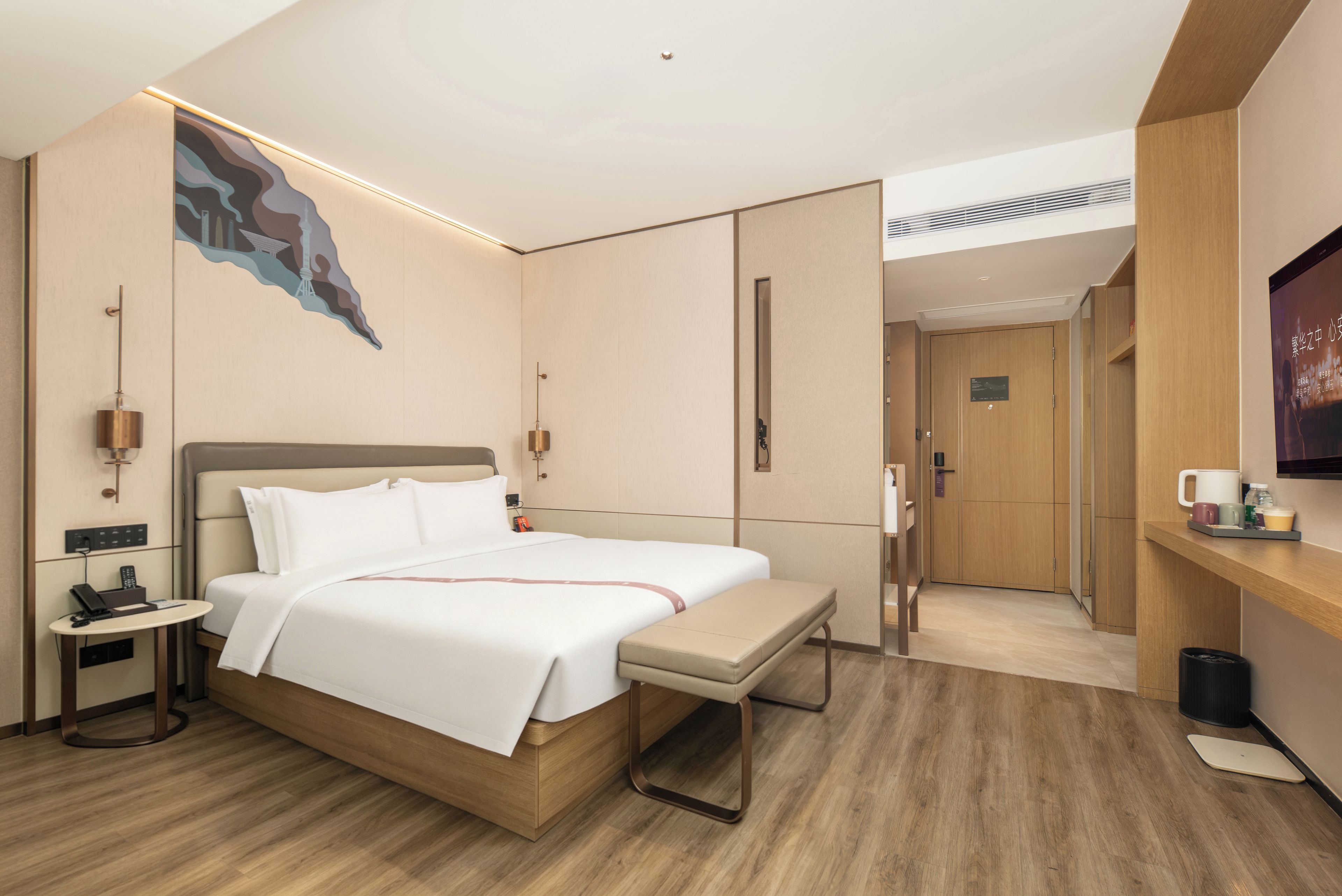 Ein modernes Hotelzimmer mit einem großen Bett, einem Nachttisch und einer Bank. An der Wand ist ein Fernseher befestigt und eine Tür führt ins Badezimmer.