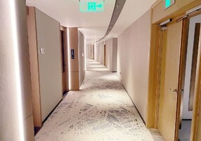 A long, narrow hallway with doors on both sides, a patterned floor, and a ceiling sign indicating an exit.