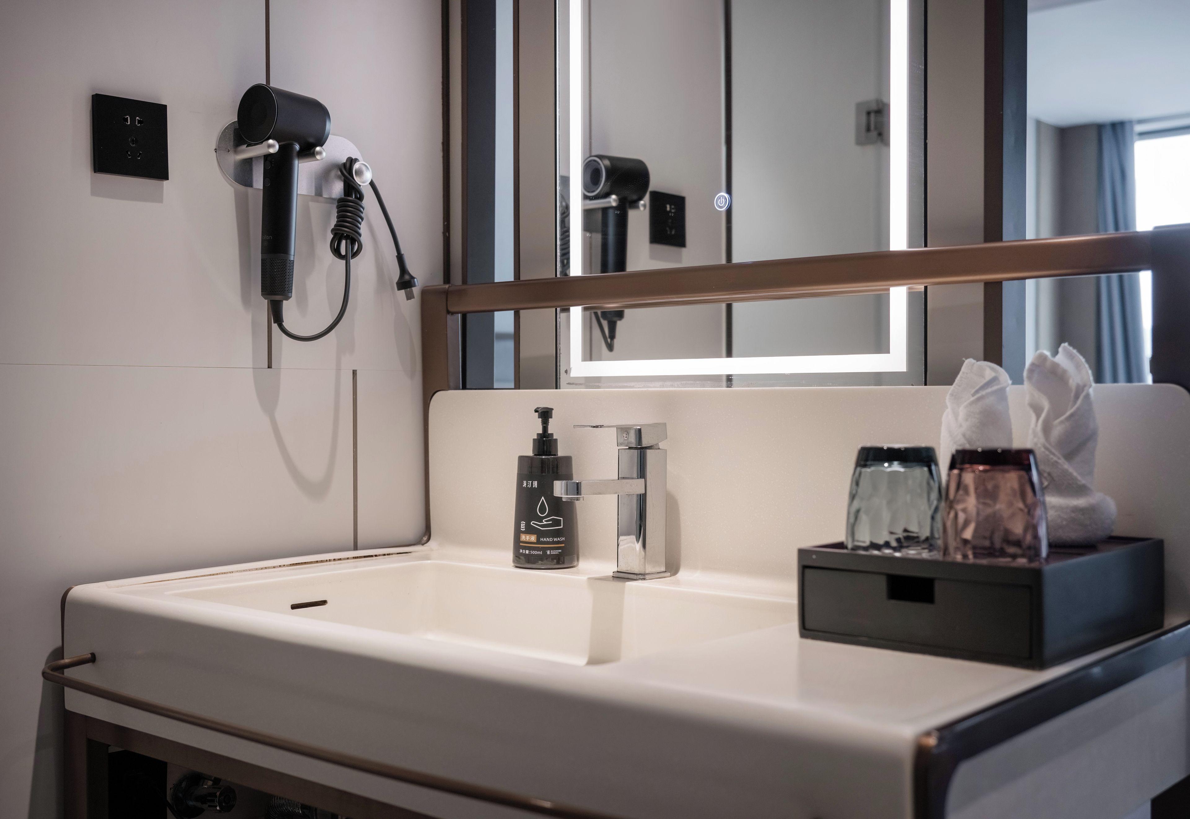 A modern bathroom with a white sink, a handheld hair dryer mounted on the wall, a bottle of hand soap, and a tissue box.