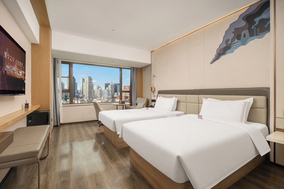 Ein Hotelzimmer mit zwei Betten, einem Schreibtisch, einem Sessel, einem Fernseher und einem großen Fenster mit Blick auf die Stadt.