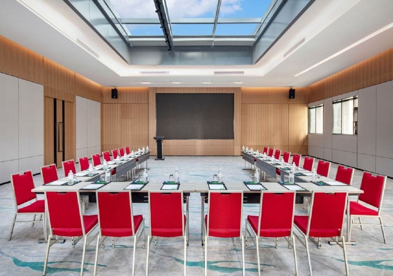A conference room with a long table, red chairs, a projector screen, and a skylight.