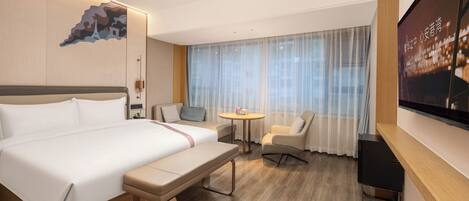 Ein Hotelzimmer mit einem Bett, einem Schreibtisch, einem Sessel, einem Fernseher und einem Fenster mit leichtem Vorhang.