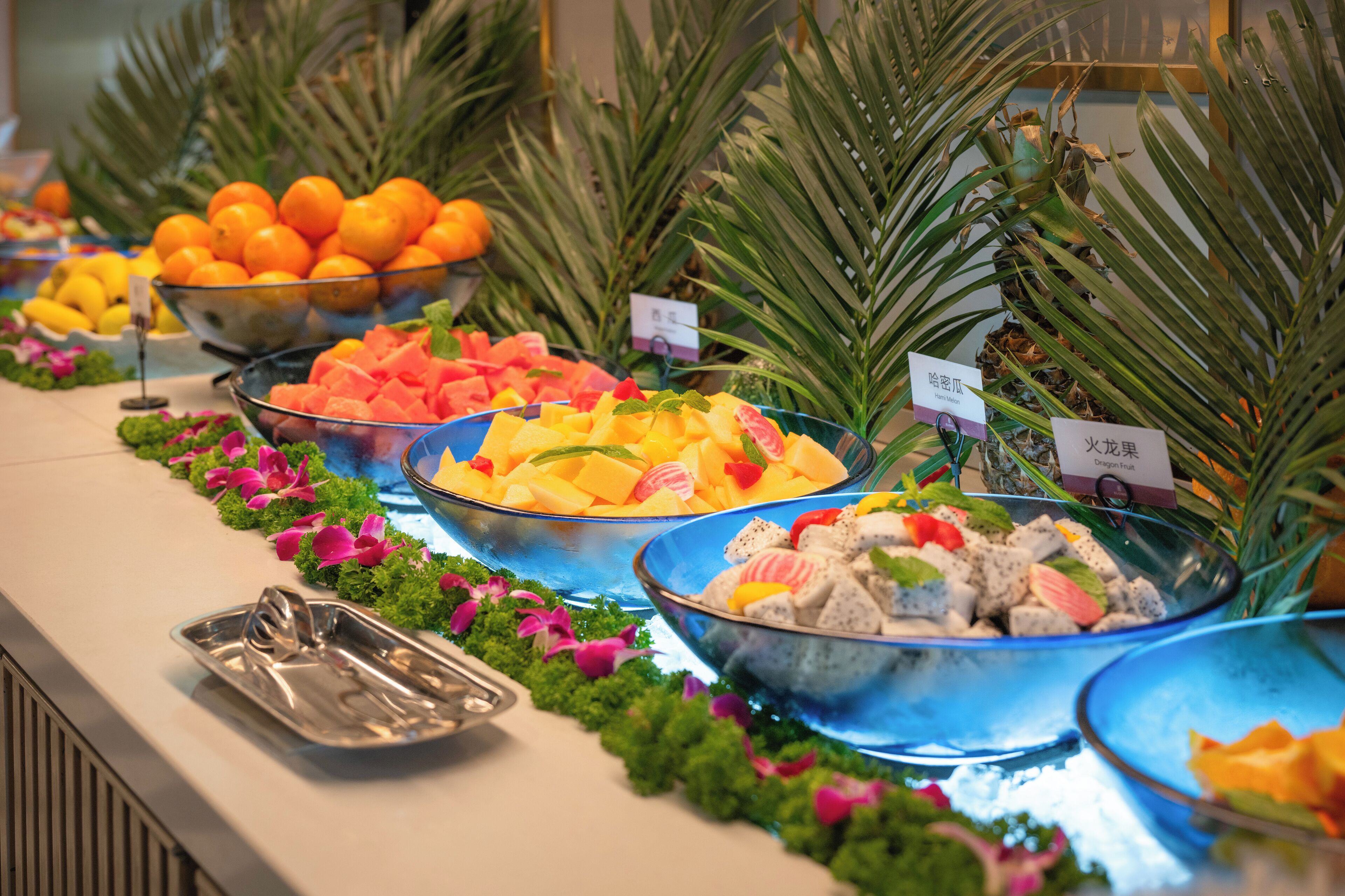 A buffet with various fruits and salads, arranged on a counter with palm plants and decorative flowers.