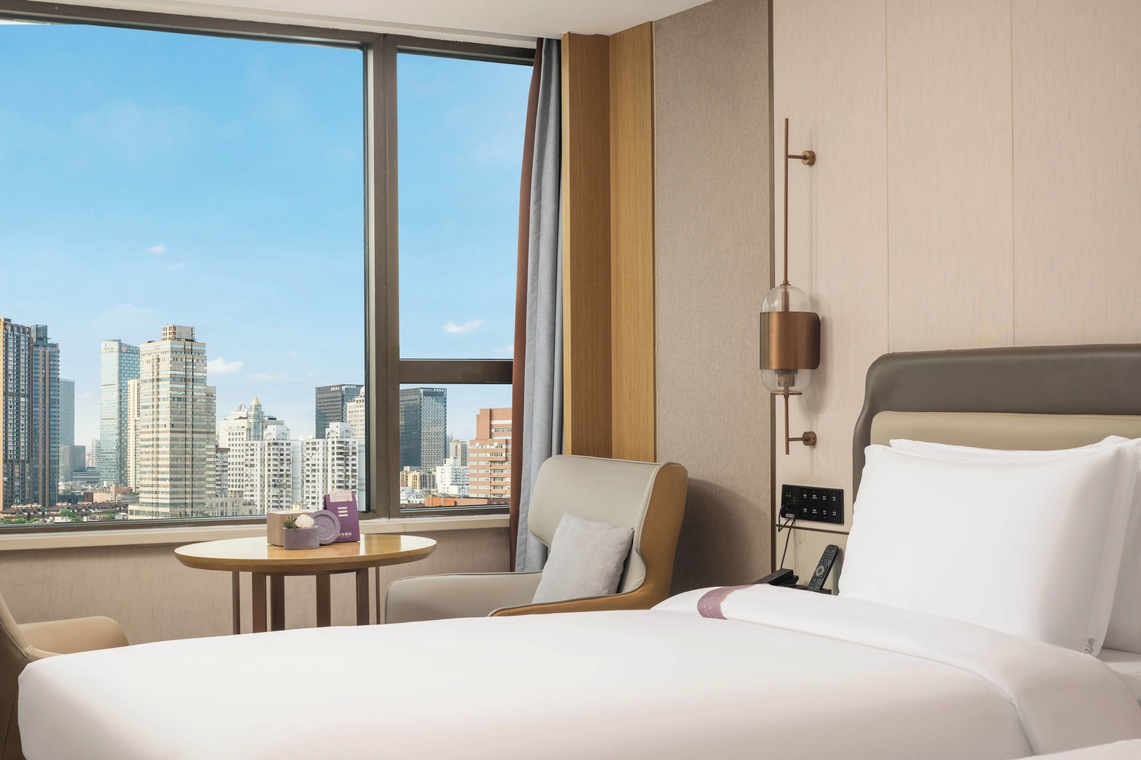 A hotel room with a large window, a bed, a chair, a small table, and a lamp.