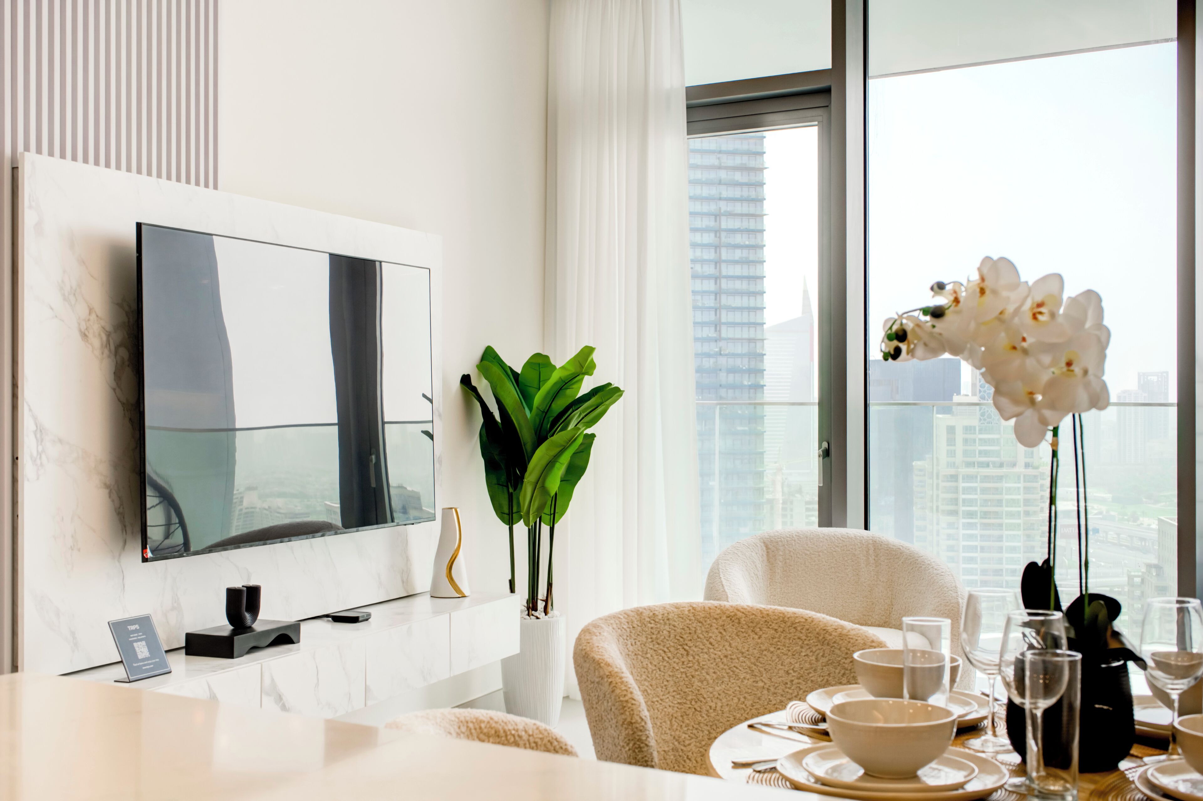 A modern hotel room with a large TV on a marble wall, a dining area with a table set for two, and a view of the cityscape through large windows.