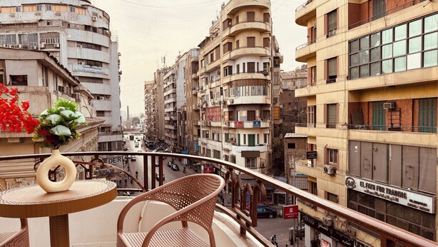 Ein Balkon mit Korbstühlen und einer Blumenvase, mit Blick auf eine belebte Straße mit mehrstöckigen Gebäuden und Geschäften.
