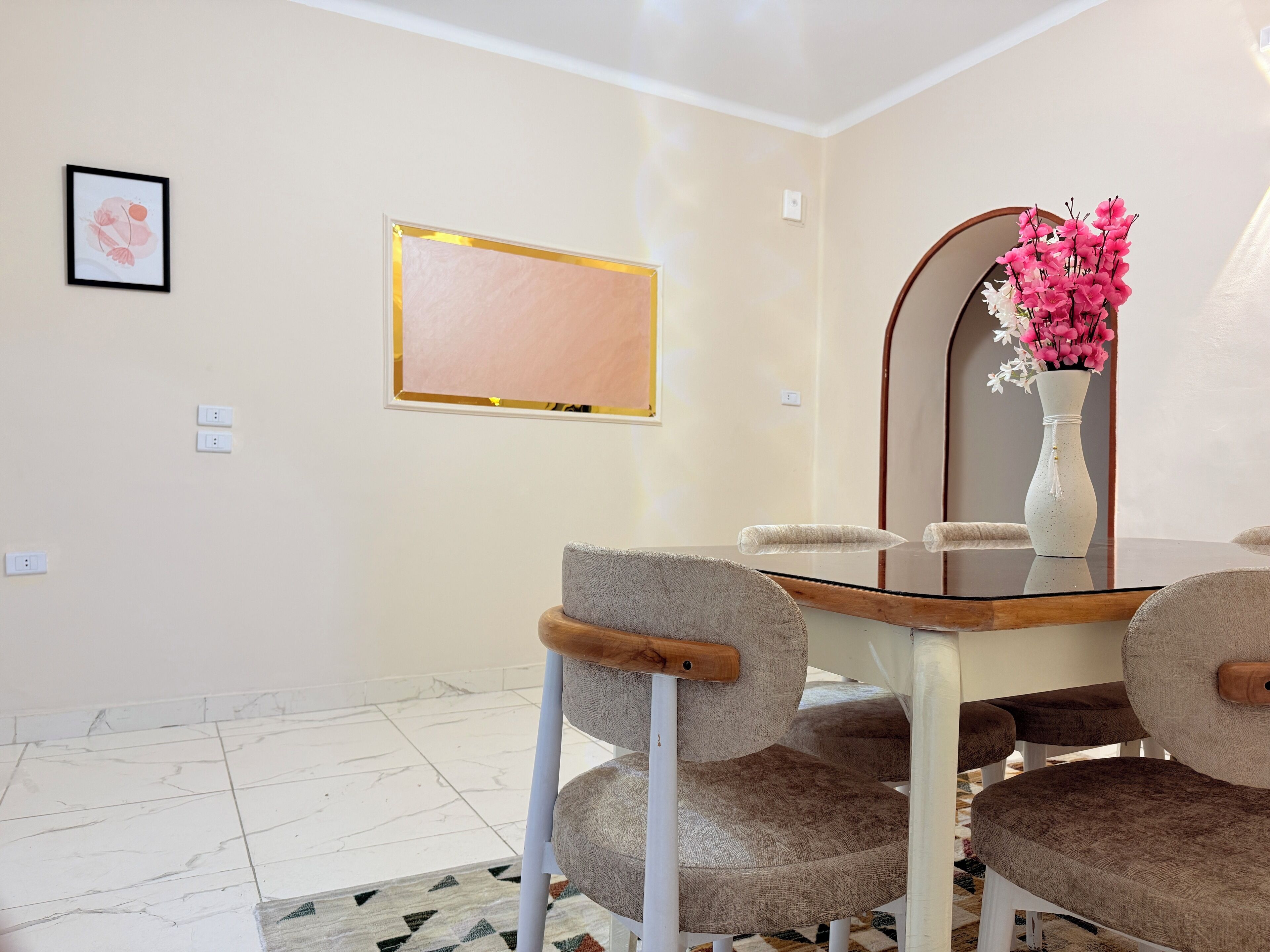 Un comedor con una mesa, sillas, un jarrón con flores, una obra de arte enmarcada y un espejo.