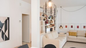 A modern living room with a white sofa, a dining table with chairs, a bookshelf, and a geometric light fixture.