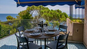 A round table set for four with chairs, a blue canopy overhead, and a view of the sea and trees.