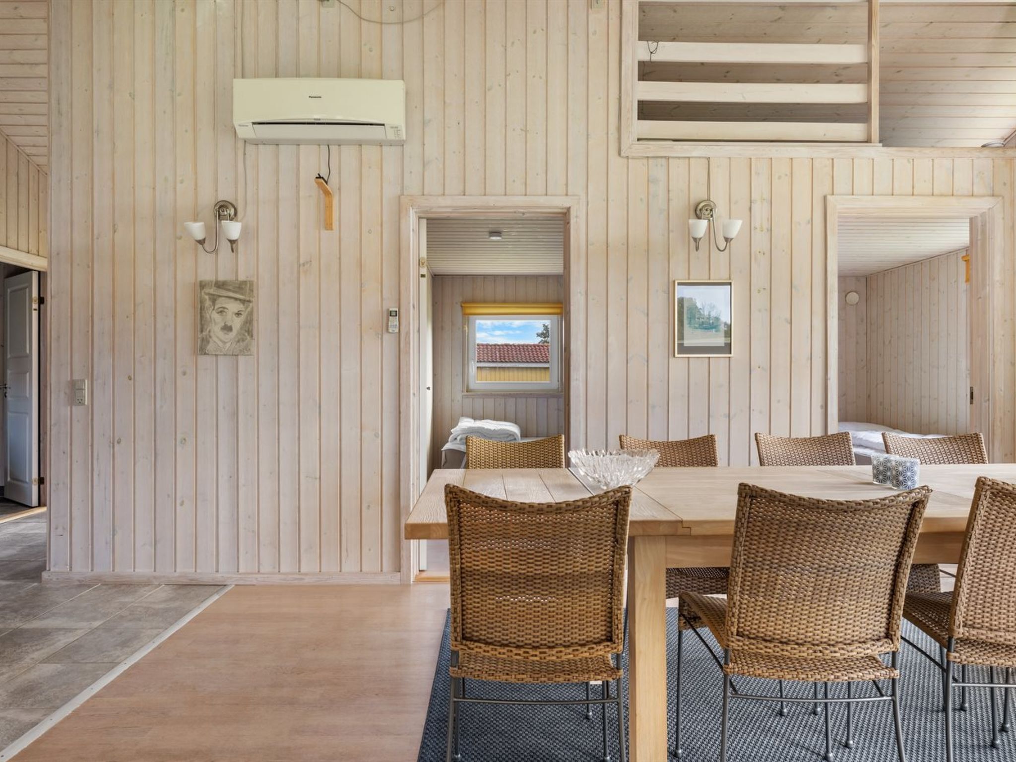 A dining area with a wooden table and wicker chairs, a wall-mounted air conditioner, and a framed picture.