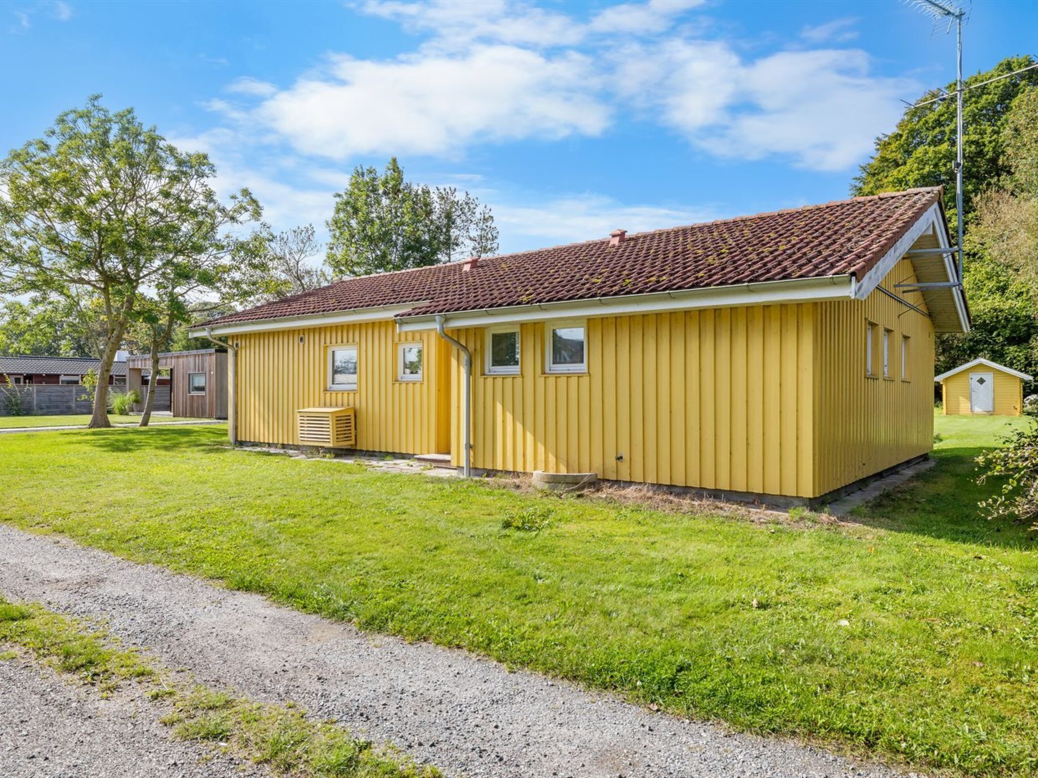 Een geel, laag gebouw met een rode pannendak, een kleine garage ernaast, en een oprit met grind.