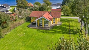 A yellow house with a red roof and a large window, surrounded by green grass and trees, with a view of the sea in the background.