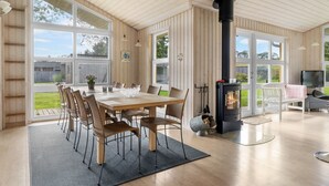 A spacious living room with a large wooden table, comfortable seating, a fireplace, and large windows offering a view of the outdoors.
