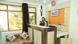 A reception area with a wooden reception desk, a cushioned bench with floral cushions, a clock on the wall, and a framed picture. - Asha Residency By St Hotel (Shimla)