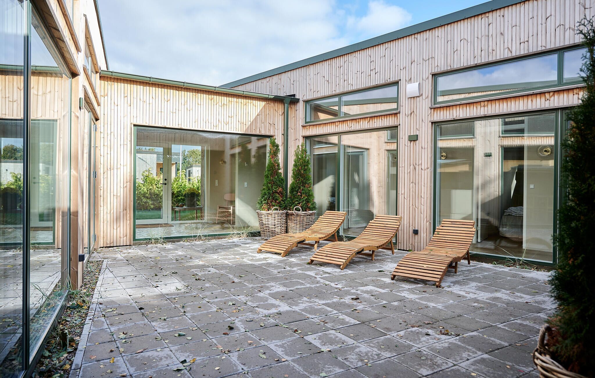 A modern building with large glass doors and windows, a paved patio with wooden lounge chairs, and potted plants.