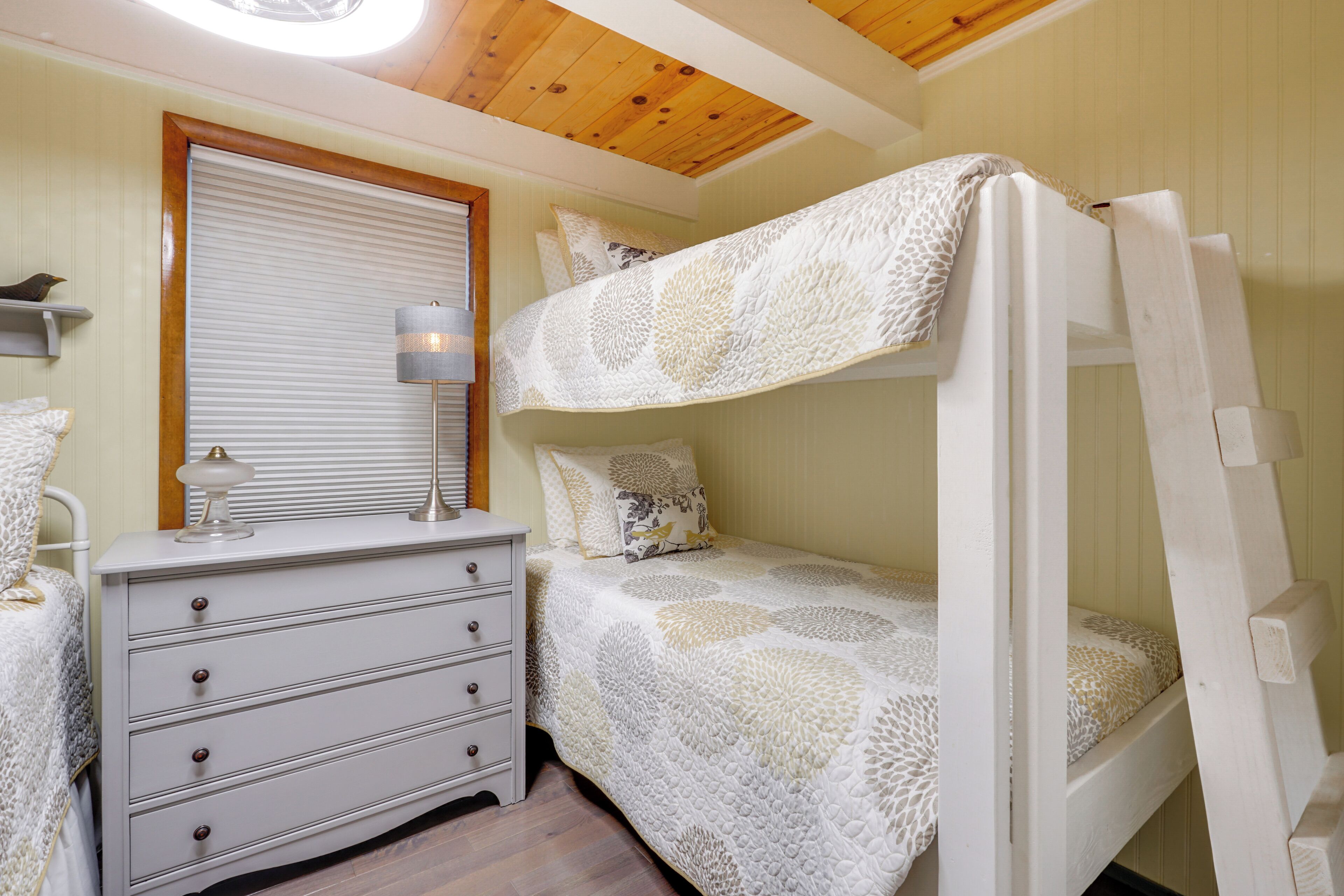 Un lit superposé avec une couvre-lit à motifs, une table de chevet avec une lampe et une fenêtre avec des stores.
