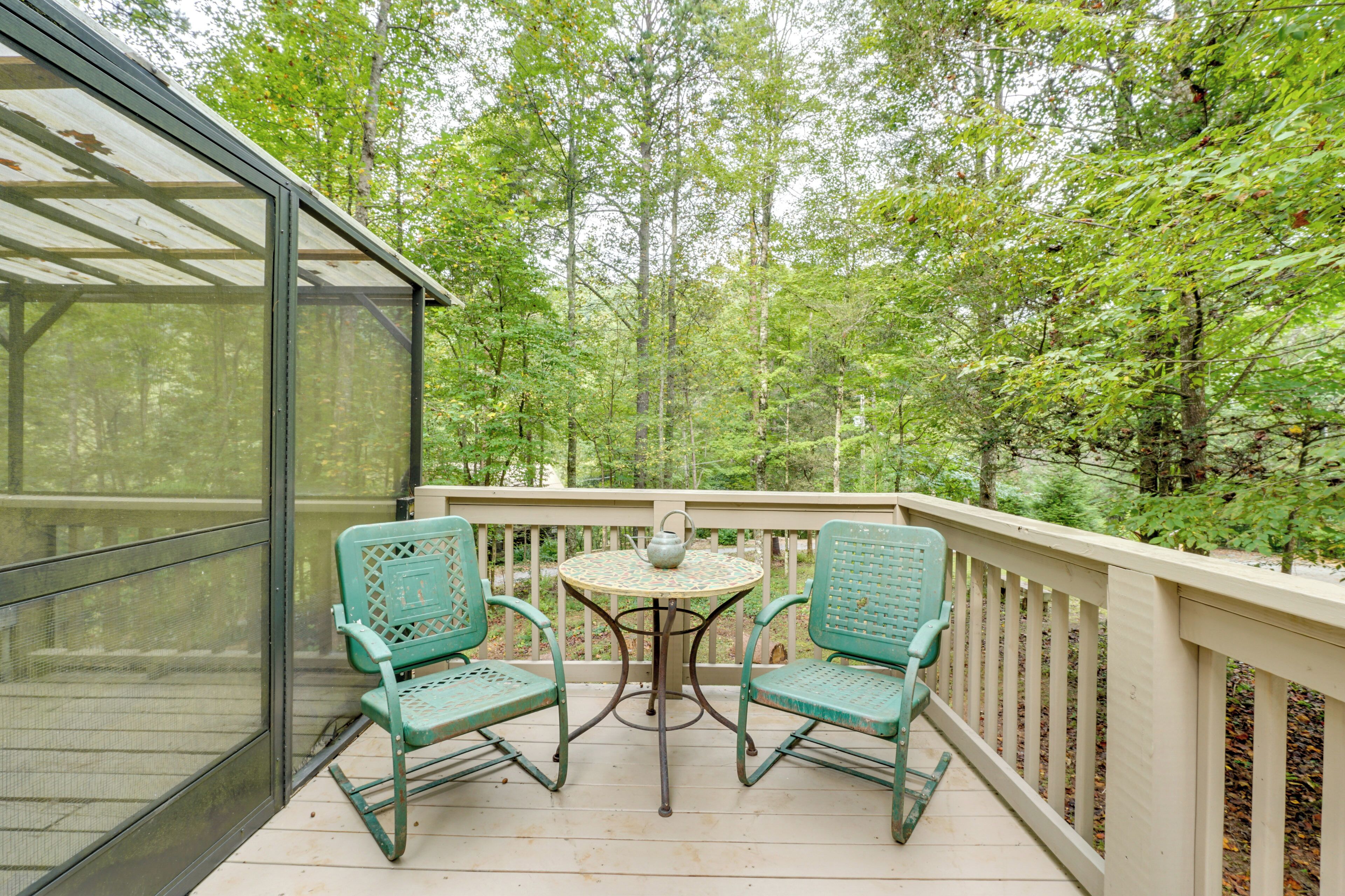A covered outdoor seating area with two chairs and a table, surrounded by a wooden deck and glass railing, overlooking a forested area.