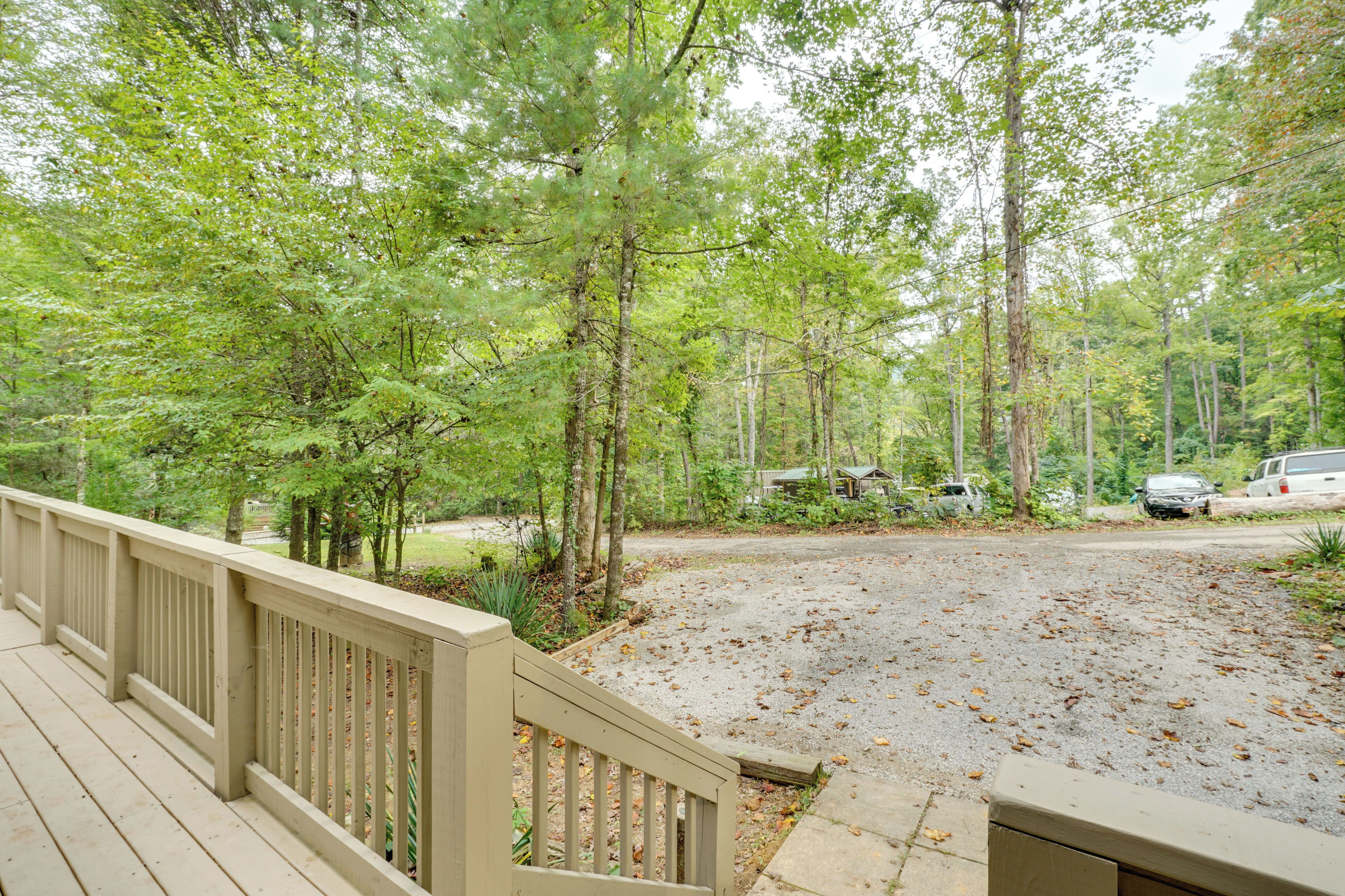 A wooden deck with a railing, surrounded by a forested area with trees and a gravel driveway.