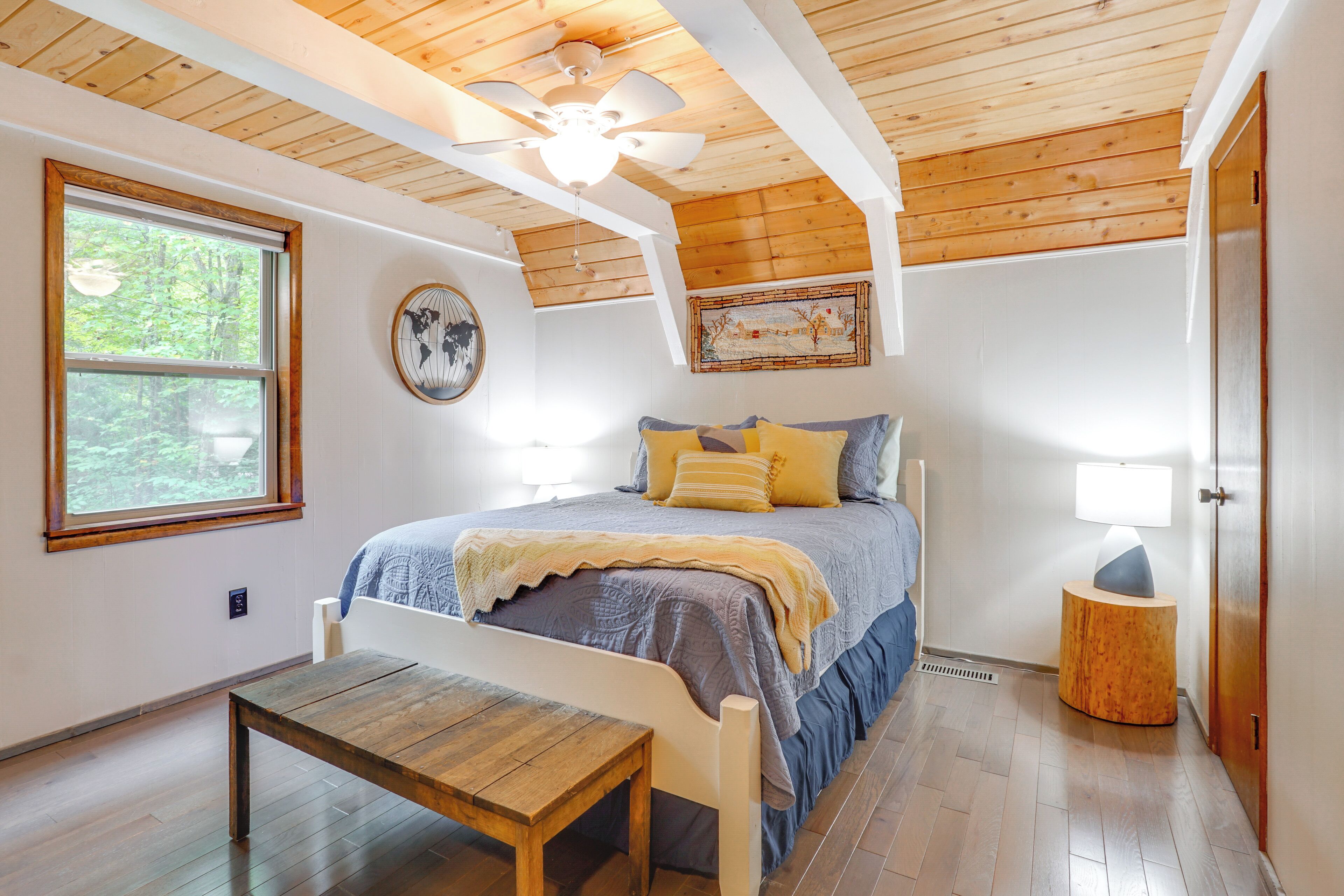 Un dormitorio con techo de madera, una cama con colcha azul y amarilla, una mesita de noche de madera, una ventana con vista a árboles y un ventilador de techo.