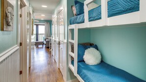 Una cama litera con ropa de cama azul, un pasillo con piso de madera y una puerta que da a otra habitación.