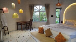 A bedroom with a large bed, a desk, and a chair. There is a window with curtains and a view of greenery outside. - Casa Rossa Resort (Pune)