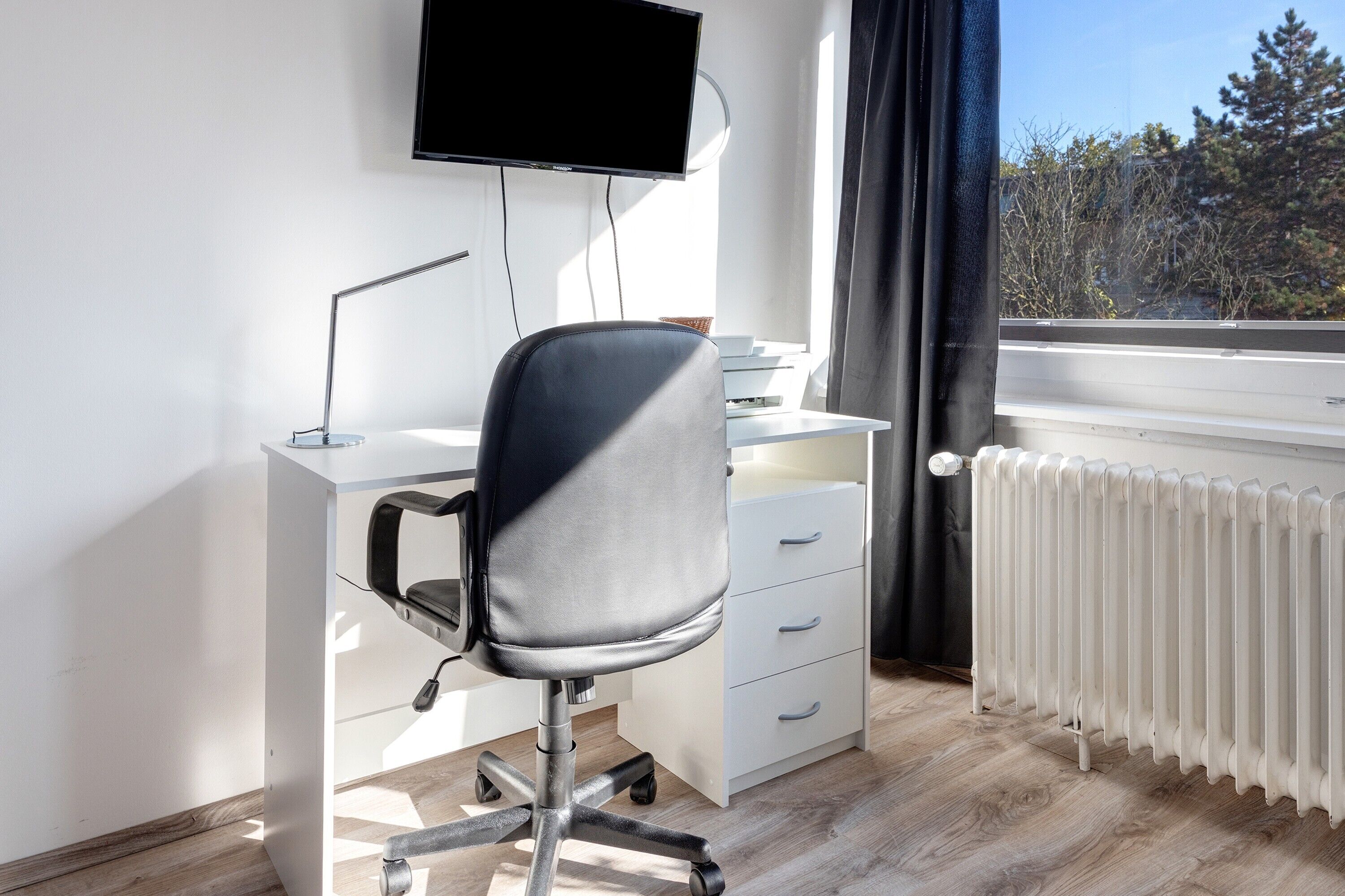 Un espacio de oficina moderno con escritorio, silla, televisor y ventana con vista a árboles.