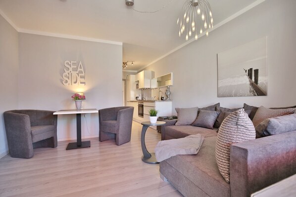 A modern living room with a sofa, two armchairs, a small table with a lamp, and a kitchen in the background.