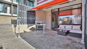 Ein moderner Außenbereich mit einer Metallstuhls und einer Couch, einem roten Sonnensegel und einem Gebäude mit großen Fenstern und einer Backsteinfassade.