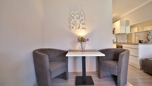 Ein modernes Wohnzimmer mit zwei grauen Sesseln, einem weißen Tisch und einer Vase mit rosa Blumen. Ein an der Wand befestigtes Schild trägt die Aufschrift "SEA SIDE".