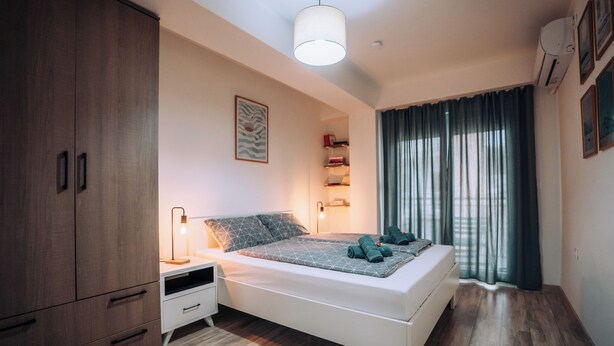 Ein modernes Schlafzimmer mit Bett, Nachttischen, einem Kleiderschrank, einer Deckenleuchte und einem Fenster mit Vorhängen.