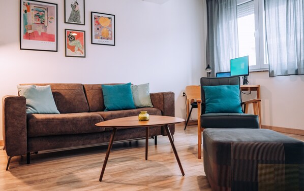 Un salon moderne avec un canapé marron, des coussins turquoise, une table basse en bois et un fauteuil noir. Des œuvres d’art encadrées ornent le mur, et un ordinateur se trouve sur un bureau.