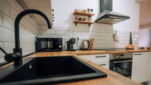 Eine moderne Küche mit einem schwarzen Spülbecken, einer Holz Arbeitsplatte, weißen Schränken, einer Mikrowelle, einem Wasserkocher und einem Regal mit Gläsern und Gewürzen.