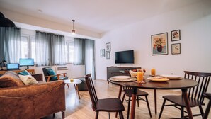 Ein modernes Wohnzimmer mit einem braunen Sofa, einem Holzesstisch und Stühlen. Ein Flachbildfernseher steht auf einem Schrank, auf einem Schreibtisch liegt ein Laptop und ein Fenster ist mit Vorhängen verziert.