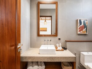 Ein Badezimmer mit Holztür, Waschbecken in Weiß, Spiegel, Handtuchhalter, Toilette und einem gerahmten Kunstwerk an der Wand.