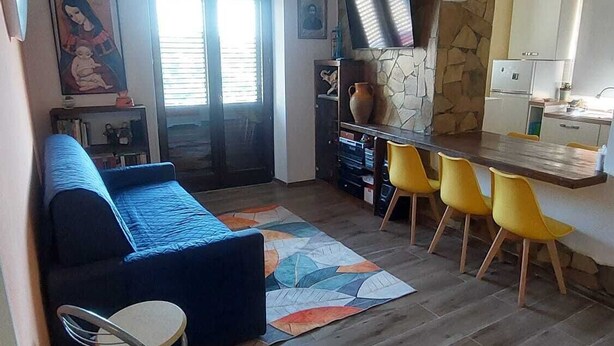 Een woonkamer met een bank, een eethoek met gele stoelen, een keuken met een stenen muur, en een raam met zonwering.