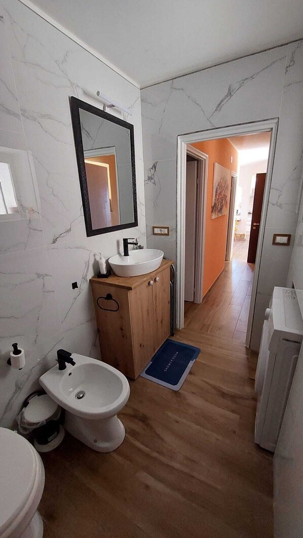 A bathroom with a marble wall, a wooden cabinet, a white sink, a bidet, and a blue shower mat.