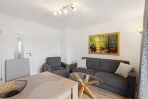 A modern living room with a glass coffee table, grey sofas, a painting of a forest, and a ceiling light fixture.