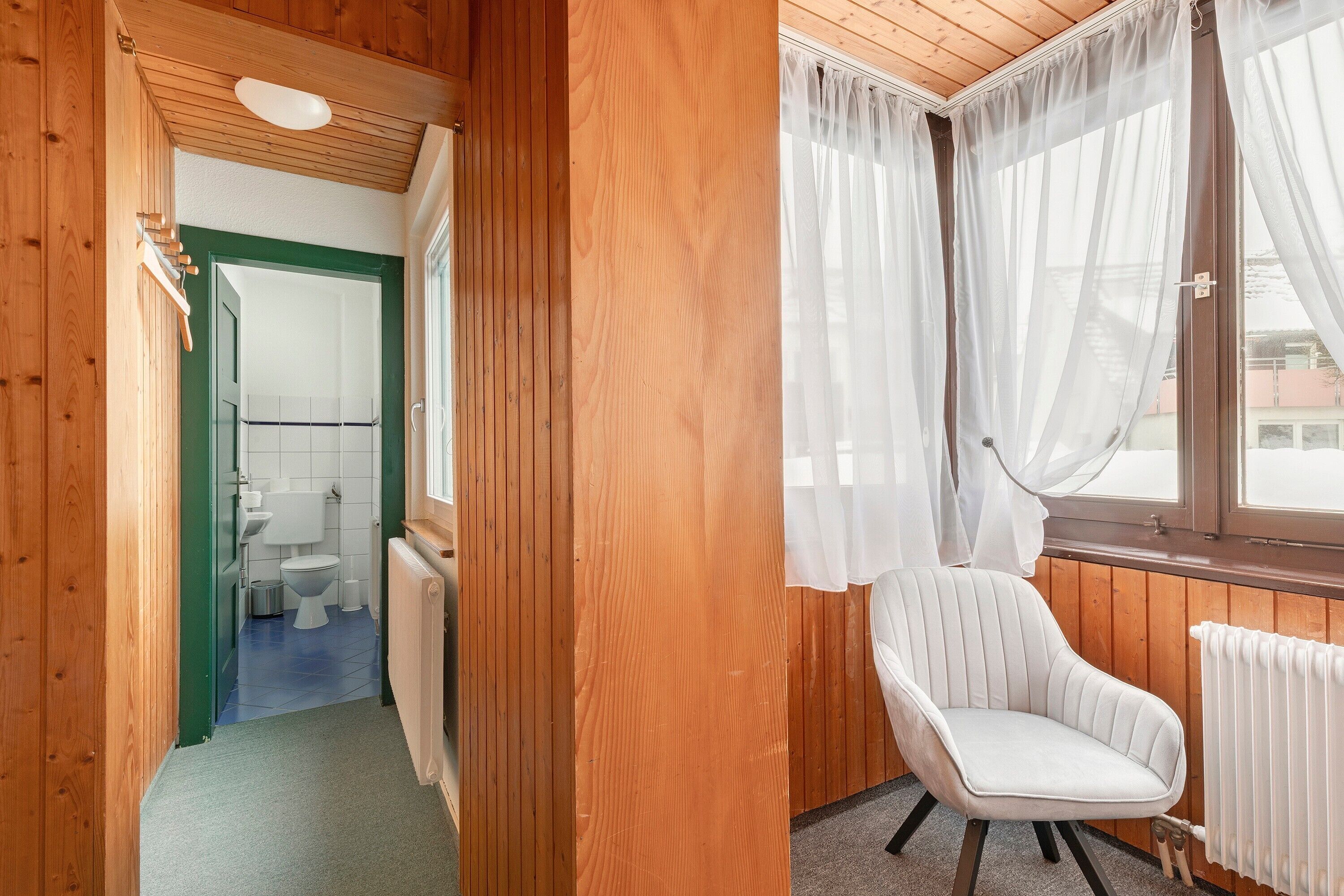 Un interior de madera con una silla blanca, una ventana con cortinas traslúcidas y vistas a un edificio.
