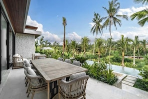 An outdoor dining area with a wooden table and chairs, surrounded by lush greenery and a pool. - Villa Maxime by BaliSuperHost (Bali)