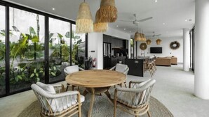 A modern interior with a round wooden table surrounded by wicker chairs, large windows with a view of greenery, and a kitchen area in the background. - Villa Maxime by BaliSuperHost (Bali)