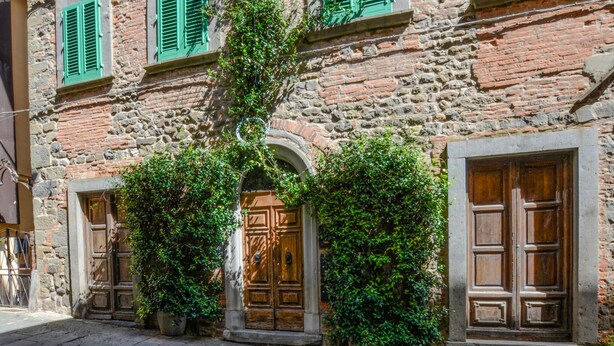 Edificio de piedra con contraventanas verdes y puertas de madera, que presenta una entrada arqueada central y plantas en macetas.