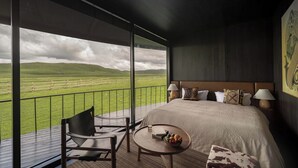 Un dormitorio con una cama grande, dos mesitas de noche con lámparas, una pequeña mesa redonda con un frutero, una silla y vistas a un paisaje verde a través de un balcón.