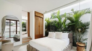 Une chambre moderne avec un grand lit, une armoire en bois, un miroir rond et une vue sur un jardin tropical, visible à travers une grande fenêtre.
