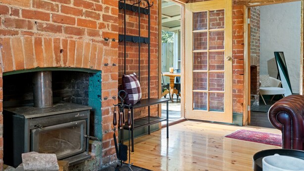 A rustic room with a brick fireplace, a wooden bench, and a door leading to an outdoor area. - Orchard Cottage—A Leafy Peaceful Retreat with Spa (Lenswood)
