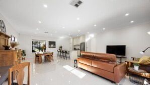 A modern living room with a brown leather sofa, a dining area with wooden furniture, a kitchen with white cabinets, and a large flat-screen TV. - Clay & Clover—Best Location Where City Meets Coast (Torrensville)