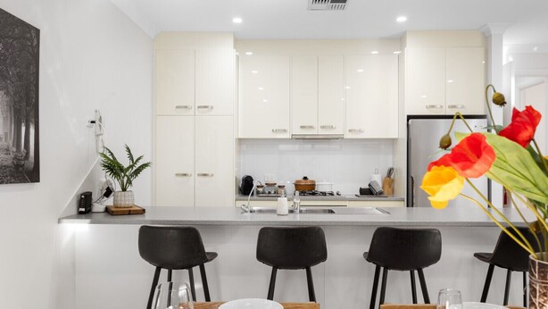 A modern kitchen with white cabinets, a central island, and black chairs. There is a dining table with white plates and glasses. - Clay & Clover—Best Location Where City Meets Coast (Torrensville)