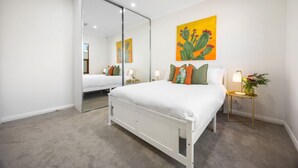 A bedroom with a large mirror, a bed with white bedding, a cactus painting on the wall, and a nightstand with a lamp and flowers. - Clay & Clover—Best Location Where City Meets Coast (Torrensville)