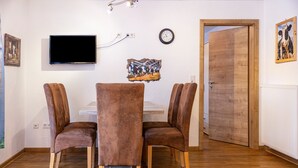 A dining area with a table and four chairs, a flat-screen TV mounted on the wall, a clock, and a wooden door.