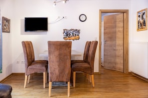 Un comedor con una mesa y cuatro sillas, un televisor de pantalla plana montado en la pared, un reloj y una puerta de madera.