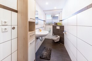 Et aflangt badeværelse med håndvask, toilet og et lille vindue.